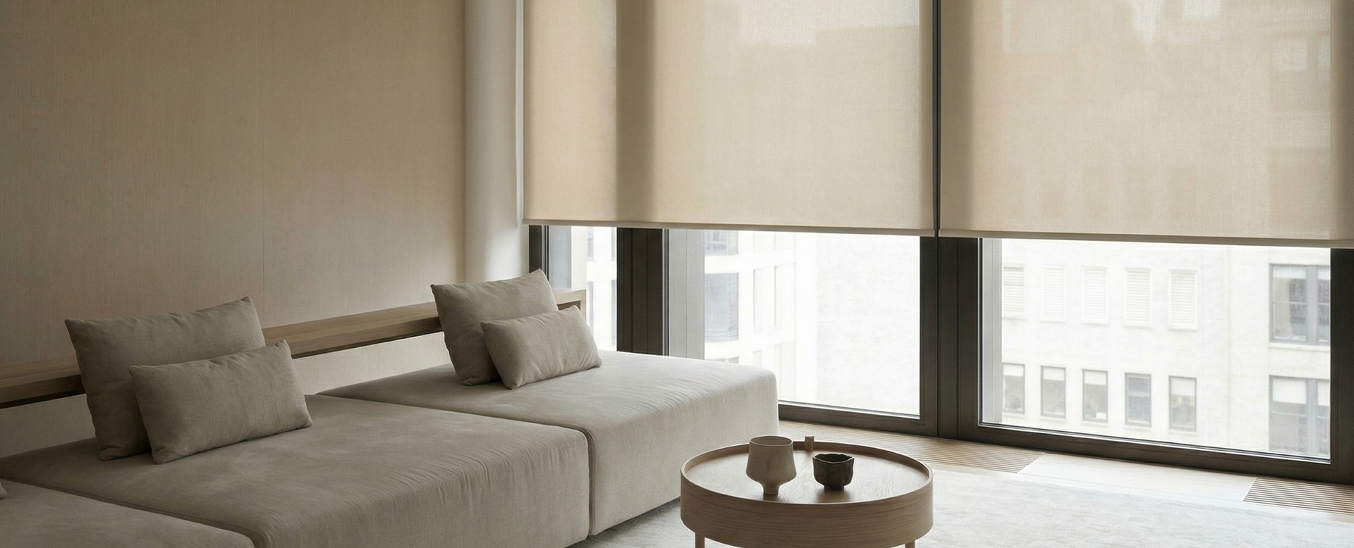 Contact The Shades Factory for a custom shades consultation while exploring this warm living room with beige roller shades on wide windows overlooking city buildings.