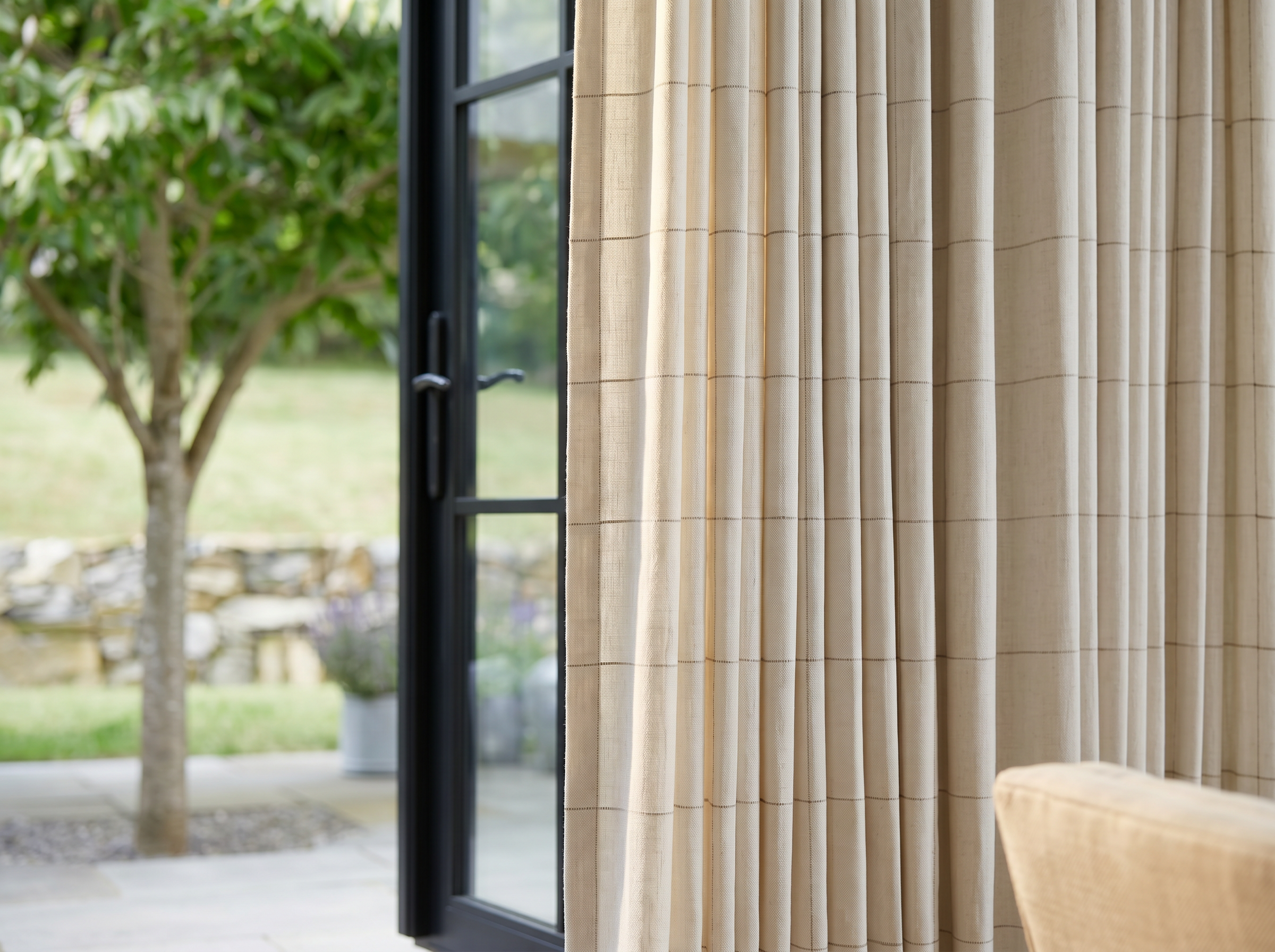 Elegant living room with floor-to-ceiling custom pleated drapes framing open black French doors overlooking a lush garden, creating a bright and inviting indoor-outdoor space.